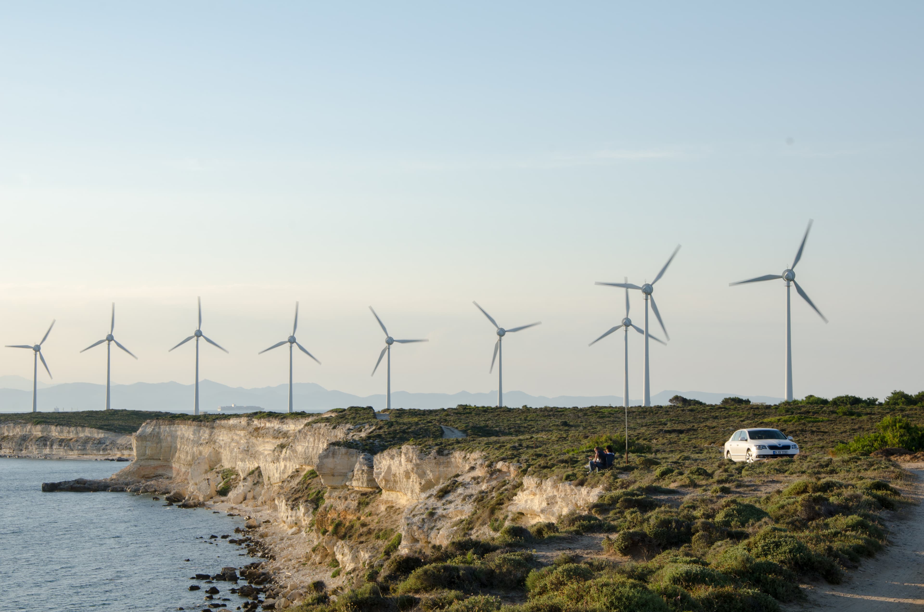 Turbine eoliche lungo una costa rocciosa della Turchia, con il mare e la vegetazione in primo piano.