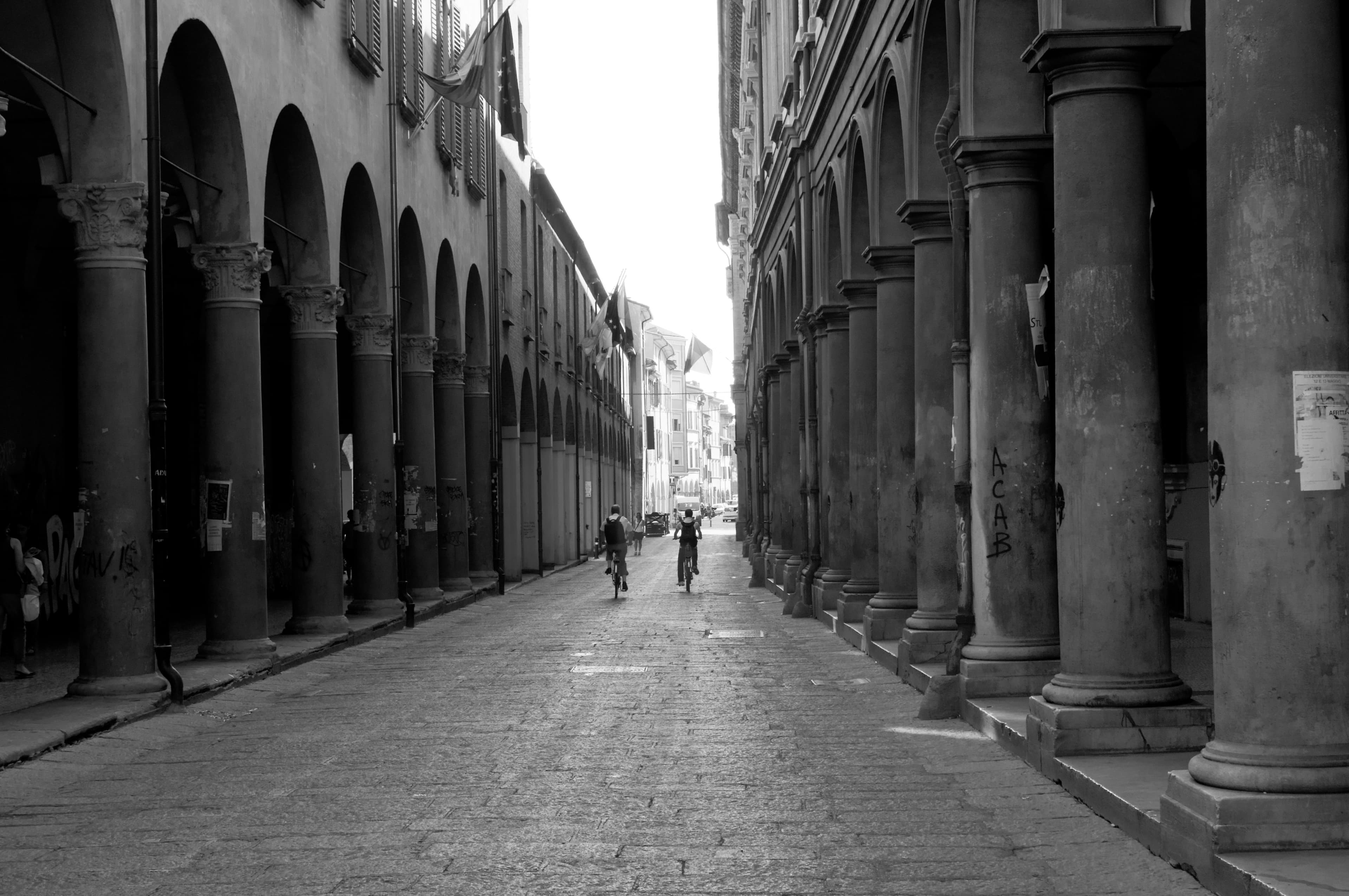 I portici di via Zamboni a Bologna, con biciclette e persone lungo la strada, in una fotografia in bianco e nero.