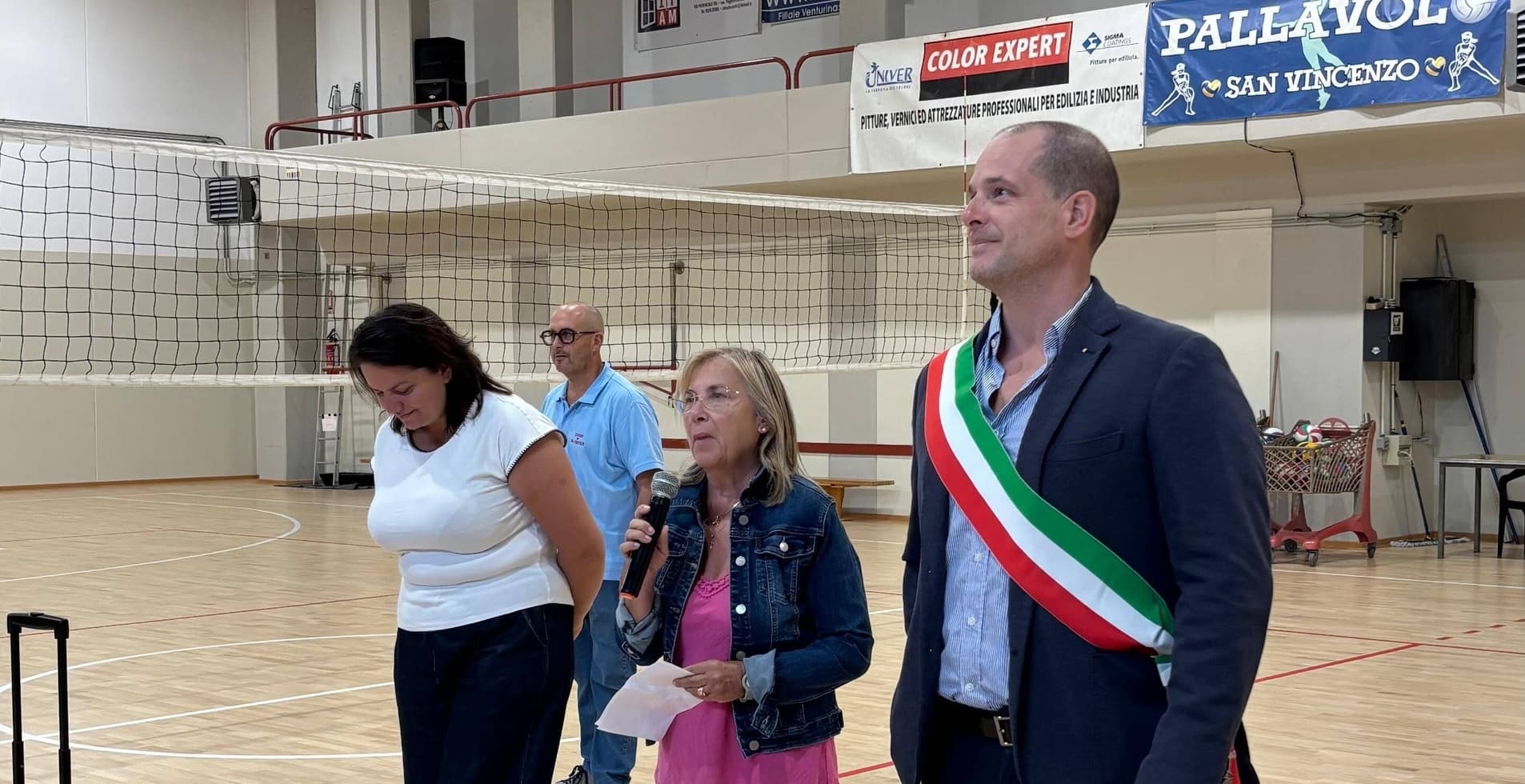 Quattro adulti alla cerimonia nella palestra Rodari, vicino a una rete da pallavolo.