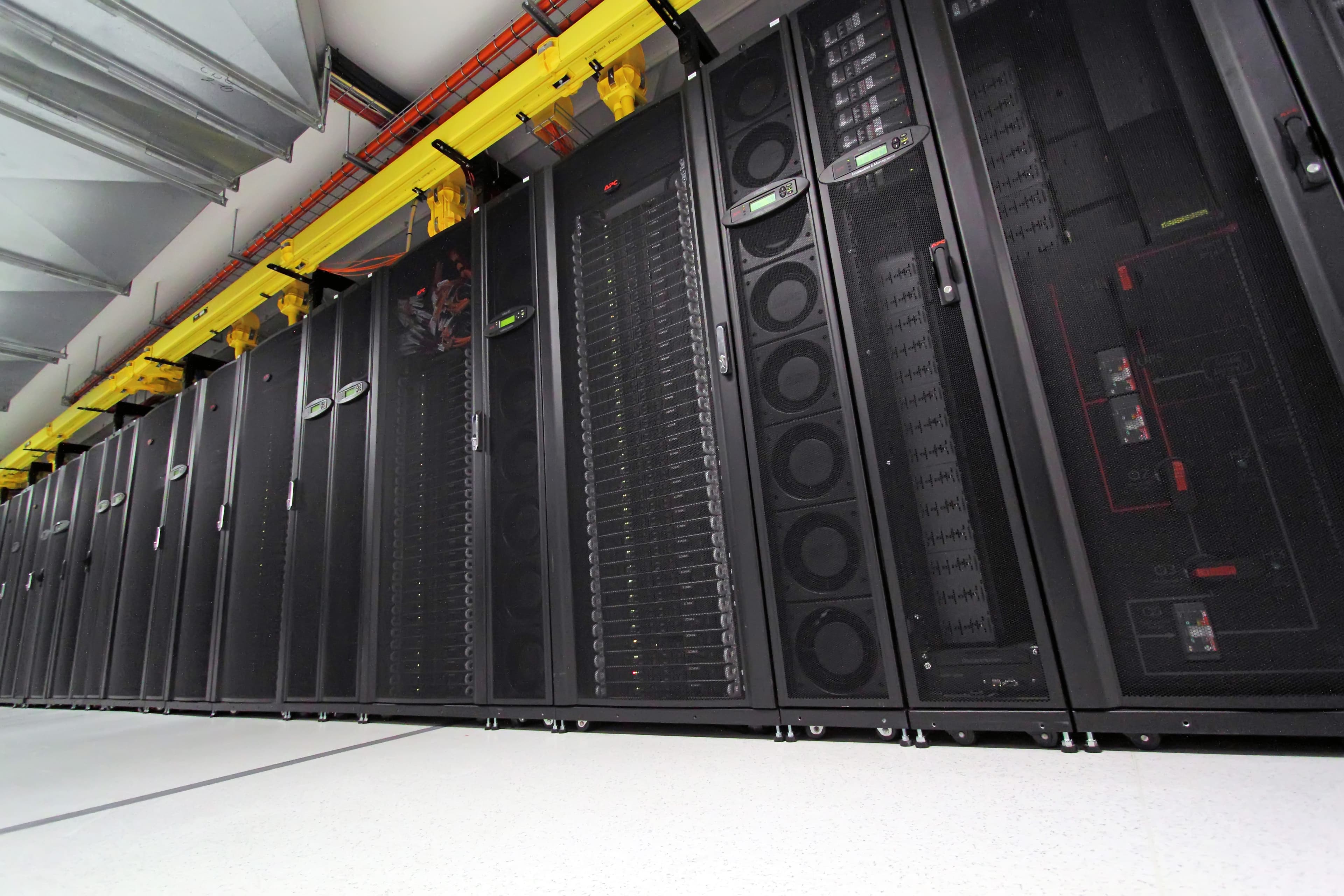 Rangée de baies informatiques noires contenant des serveurs, avec des câbles jaunes au-dessus.