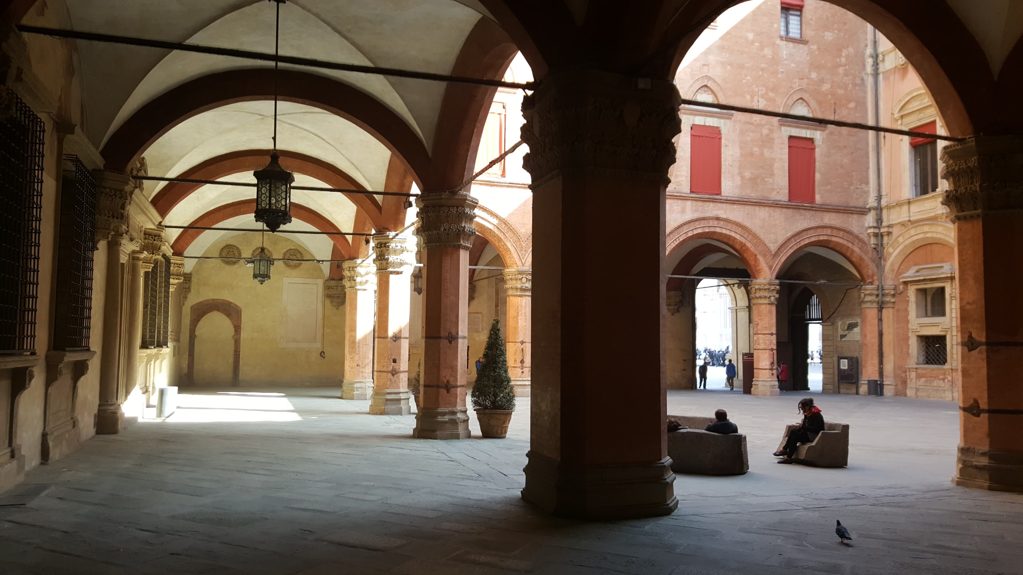 Archi e colonne del cortile di Palazzo d’Accursio a Bologna, con alcune persone sedute sulle panchine.