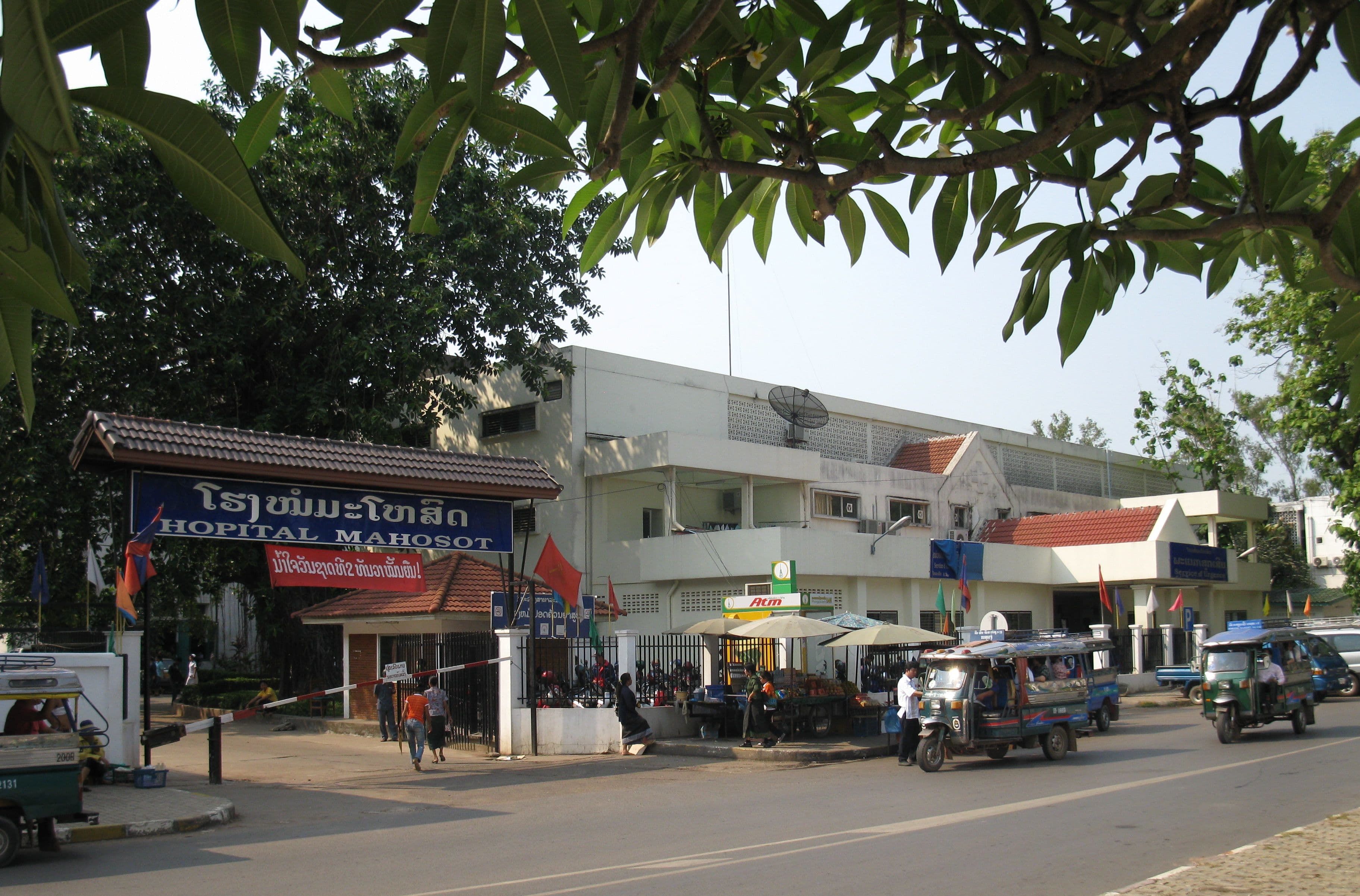 Ingresso dell’ospedale Mahosot a Vientiane, con alberi e veicoli sulla strada