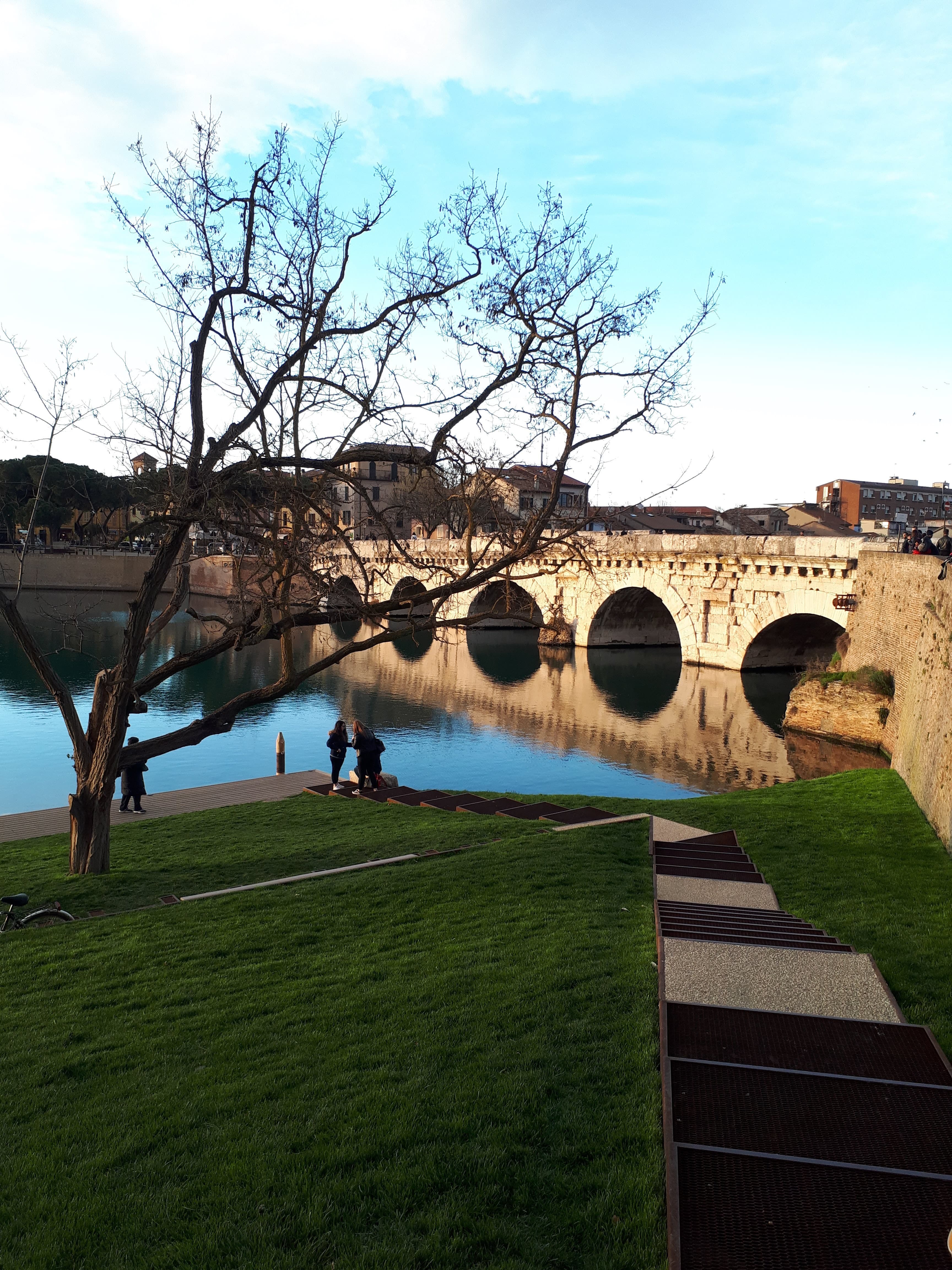 Il ponte di Tiberio a Rimini, con alberi, un prato e un camminamento lungo l’acqua.